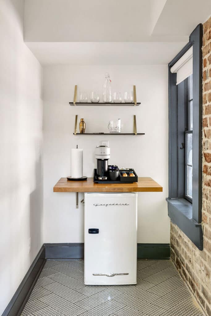 Compact coffee corner with a white mini fridge, wooden counter, and shelves of glassware by a window.
