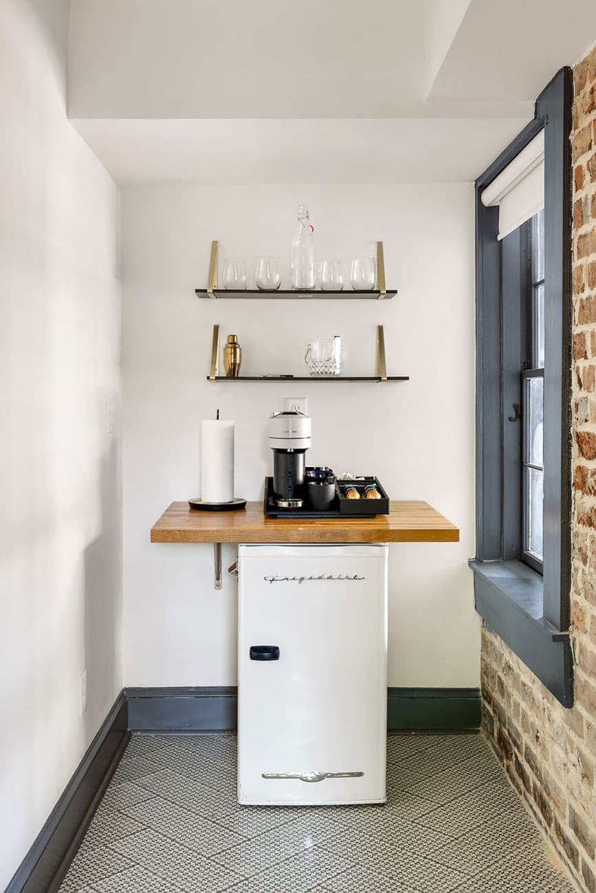 Compact coffee corner with a white mini fridge, wooden counter, and shelves of glassware by a window.