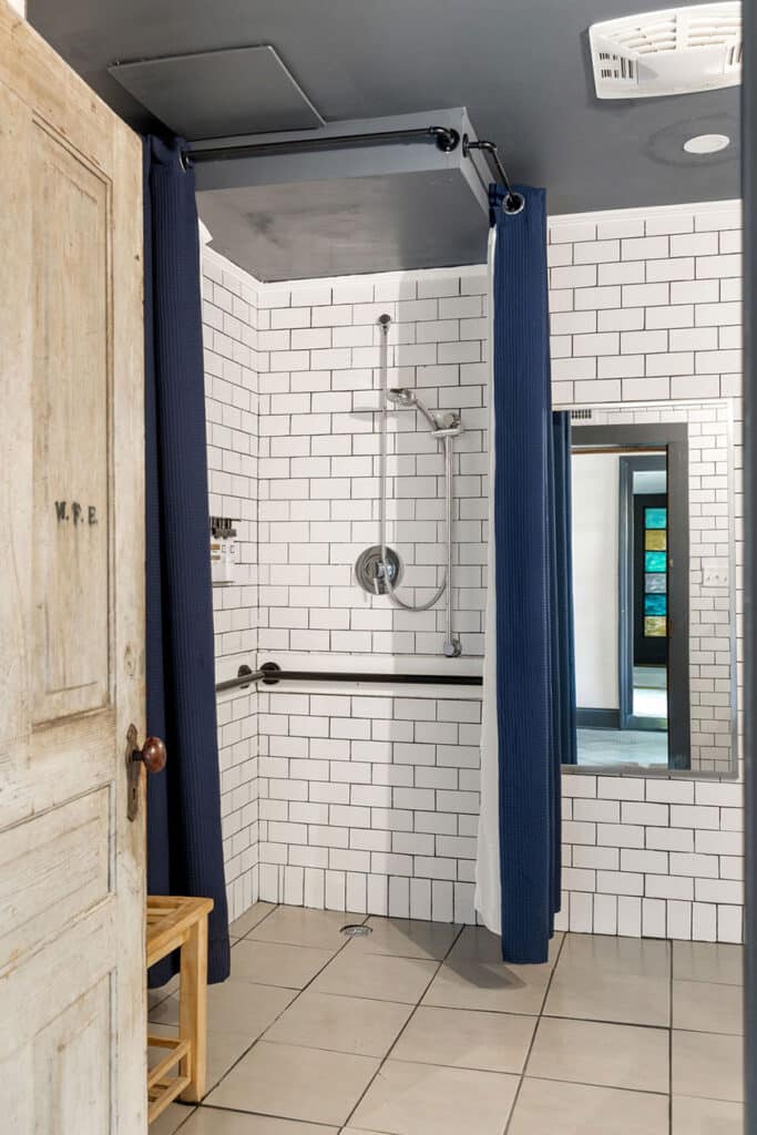 Shower stall with white subway tiles, blue curtain, handheld showerhead, and gray floor tiles; small wooden bench to the left.