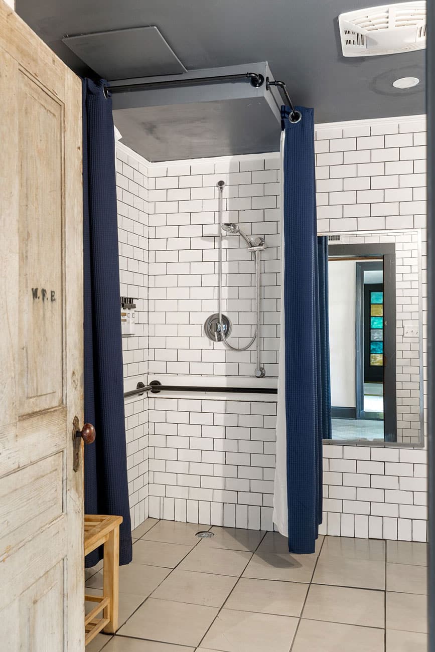 Shower stall with white subway tiles, blue curtain, handheld showerhead, and gray floor tiles; small wooden bench to the left.