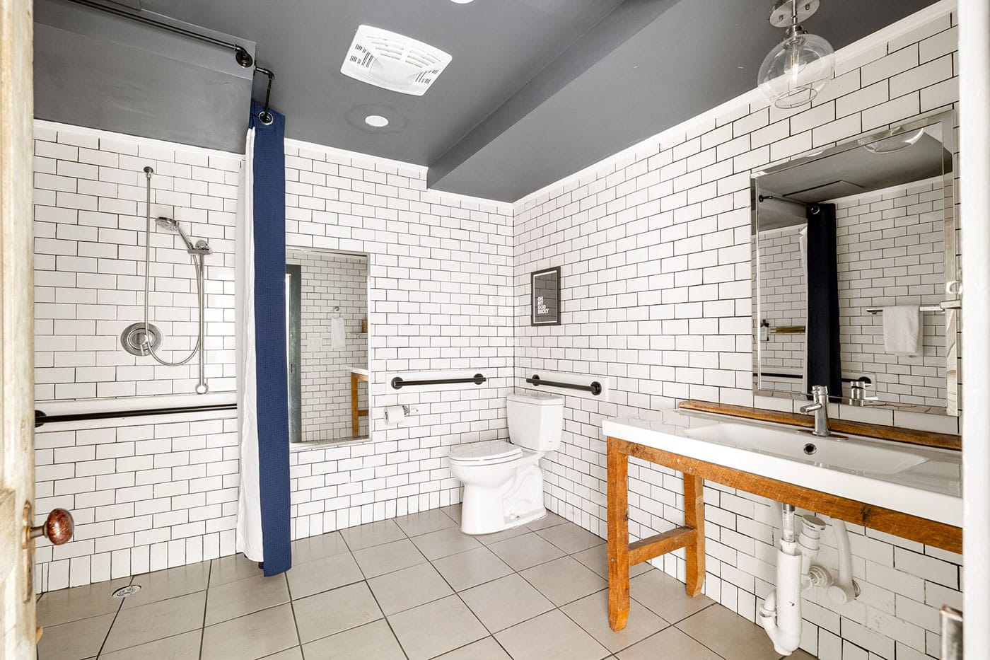 Accessible bathroom with white subway-tiled walls, a walk-in shower with a blue curtain, and a toilet with wall grab bars, plus a wooden-framed sink on the right.