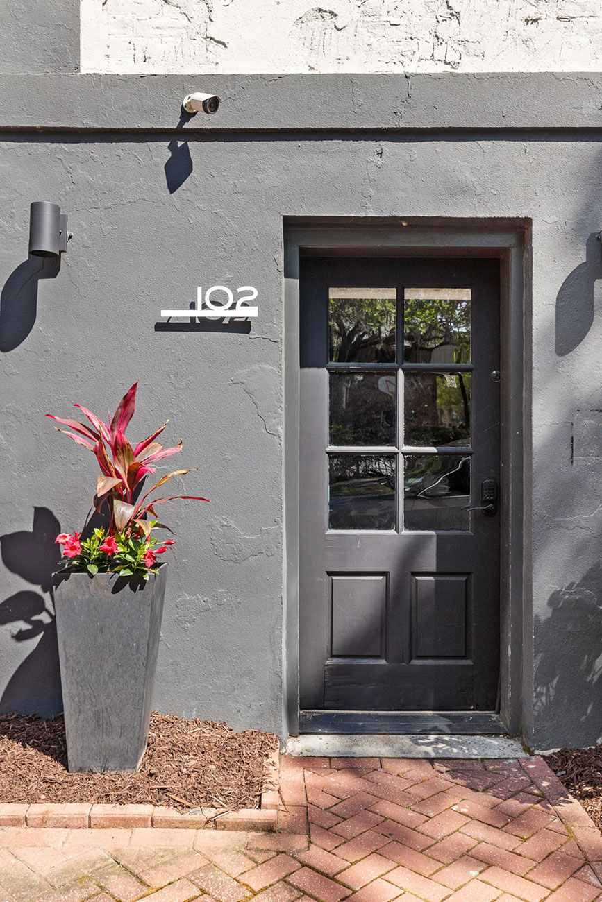 Gray building entrance with a black pane door, a tall planter of red plants to the left, the number 102 on the wall, and a security camera above.