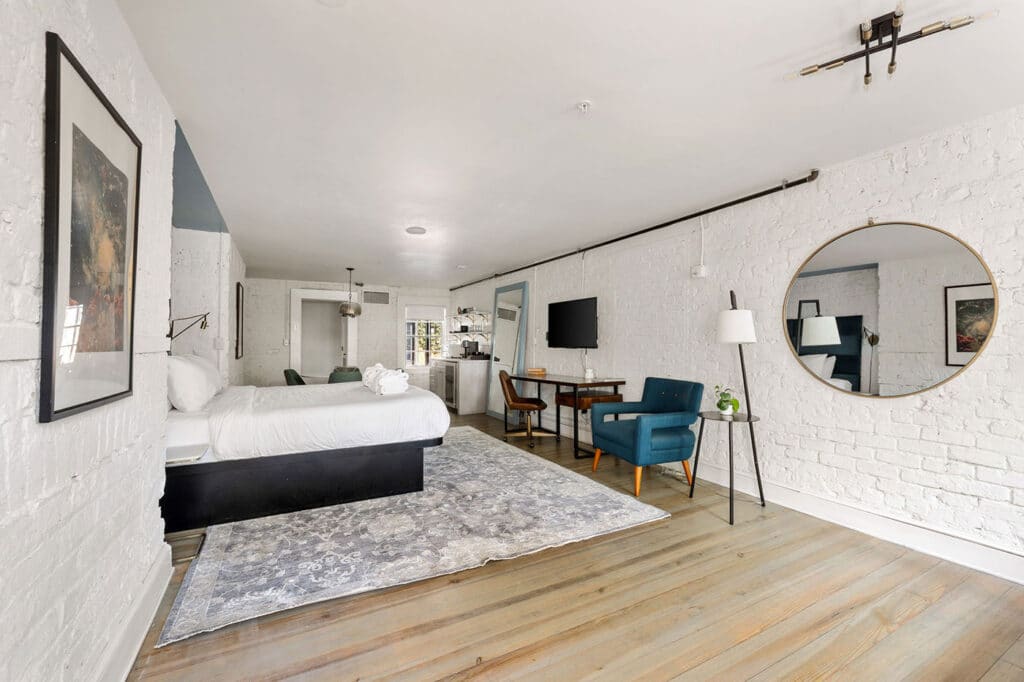Bright loft-style hotel room with a white brick wall, a large bed, and a blue chair beside a round mirror and small desk area.