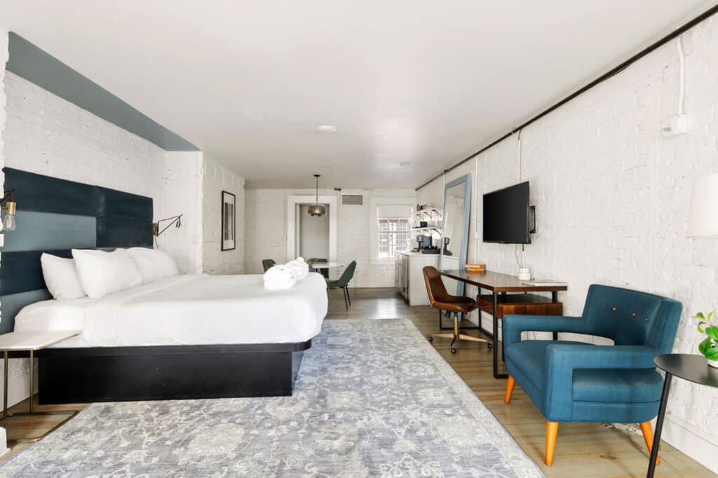 Room with white brick walls, king bed with a dark blue headboard, plush pillows, and a light gray patterned rug. A teal chair and desk area sit along the wall, with a mounted TV nearby.