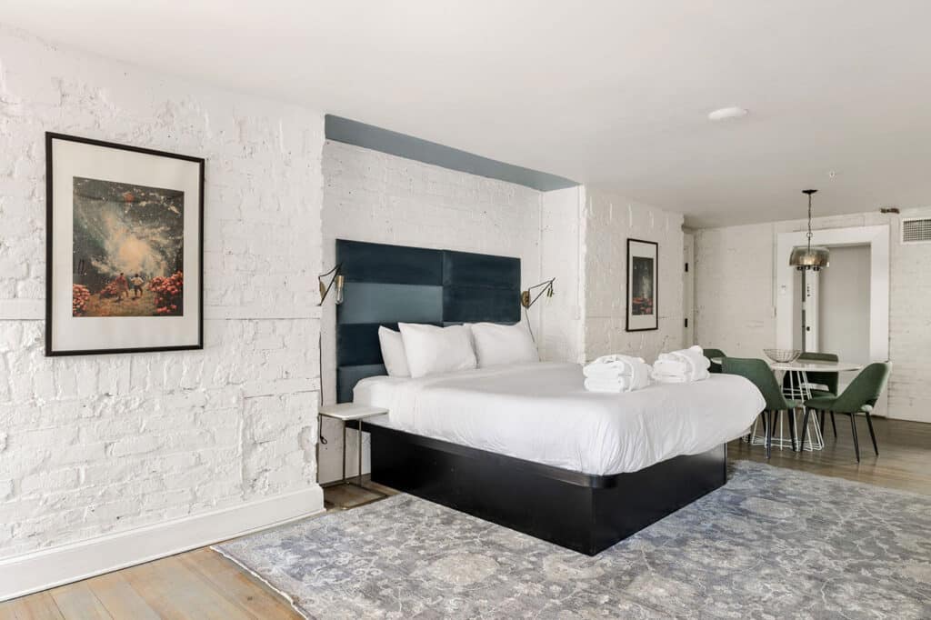 Hotel room with white brick walls and a dark blue headboard, a king bed with white linens and folded towels, two brass wall lamps, and a small seating area with green chairs and a round table on the right.