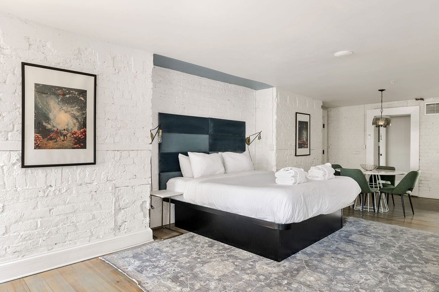 Hotel room with white brick walls and a dark blue headboard, a king bed with white linens and folded towels, two brass wall lamps, and a small seating area with green chairs and a round table on the right.