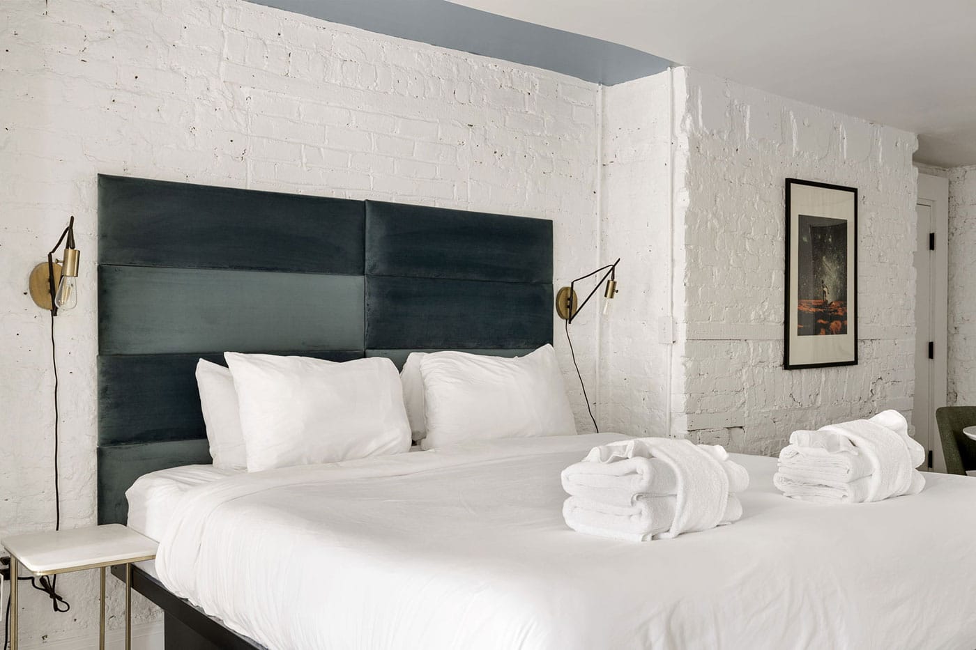 White-brick bedroom with a dark blue padded headboard, white bedding, and two folded towels on the bed.