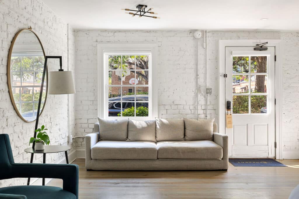 Bright living room with a light gray sofa, teal chair, round wall mirror, and white brick walls bathed in daylight from windows and a glass-pane door.