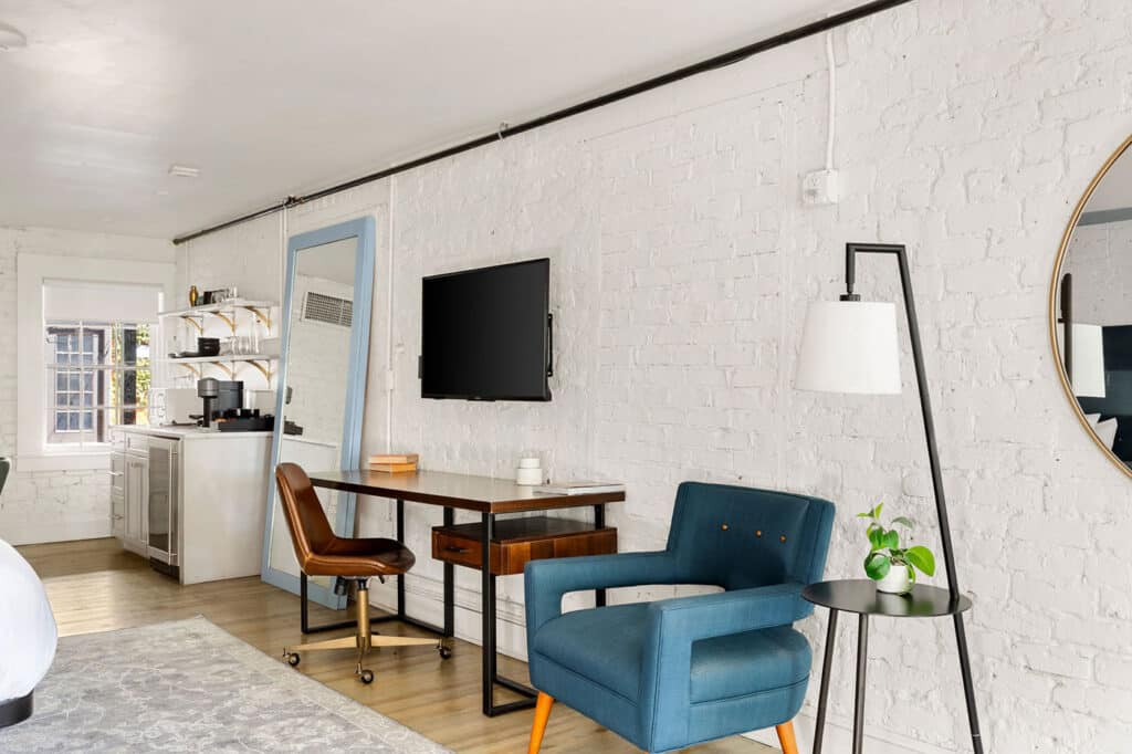 Bright loft living/working space with a blue armchair, desk and chair, wall-mounted TV, floor lamp, plant on a small side table, and a round mirror on white brick wall