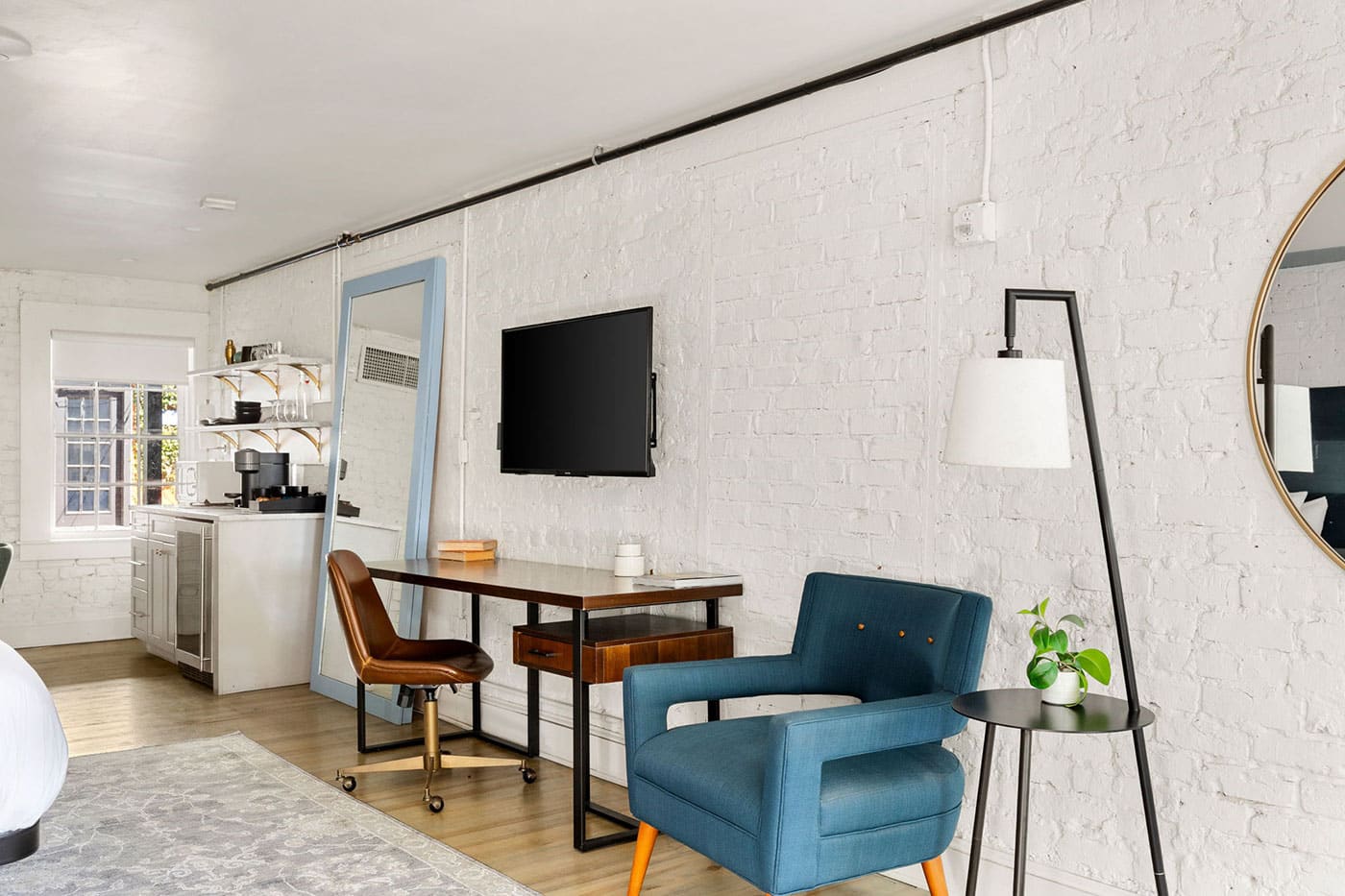 Bright loft living/working space with a blue armchair, desk and chair, wall-mounted TV, floor lamp, plant on a small side table, and a round mirror on white brick wall