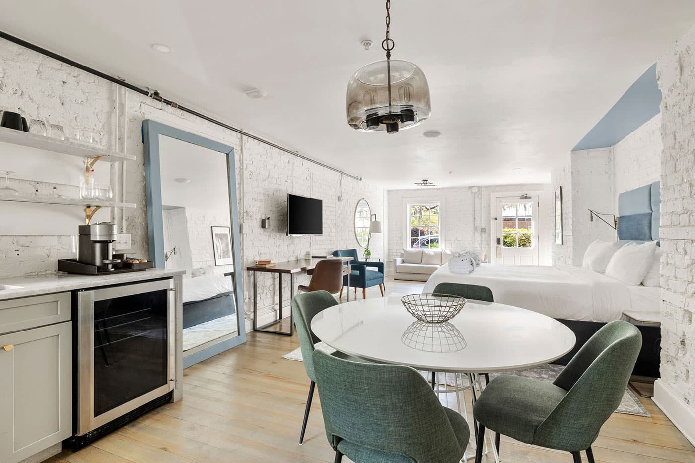 Bright open-concept studio with white brick walls, round white dining table, and green upholstered chairs; bed and living area visible in the background.