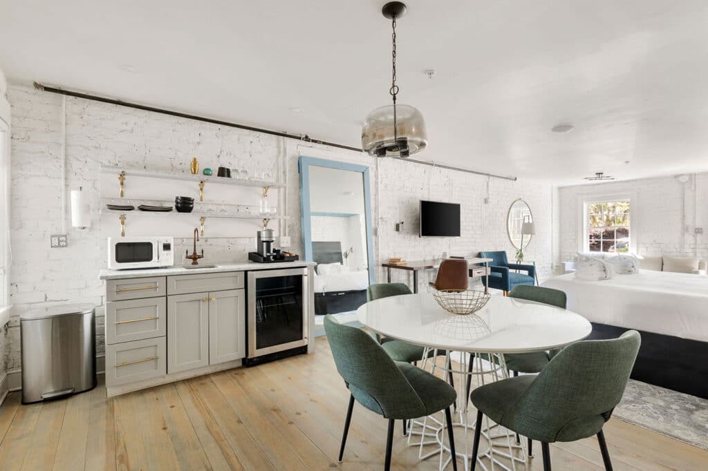 Open-plan studio with a kitchenette, round white dining table, and green chairs near a sleeping area against white brick walls.
