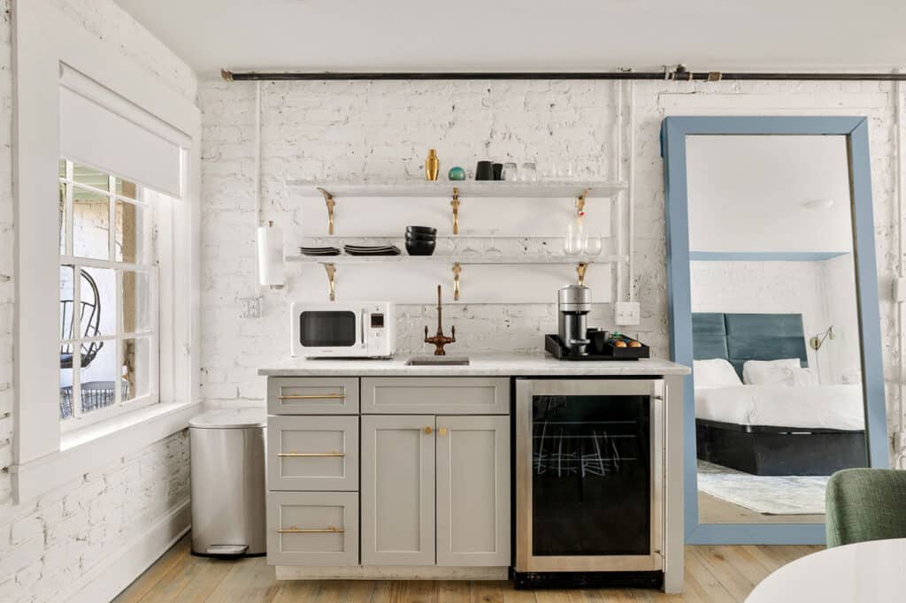 Loft-style kitchenette with white brick walls, gray cabinets, brass handles, and open shelves holding dishes, a microwave, and a coffee station; a large blue-framed mirror reflects a bedroom with a bed in the background