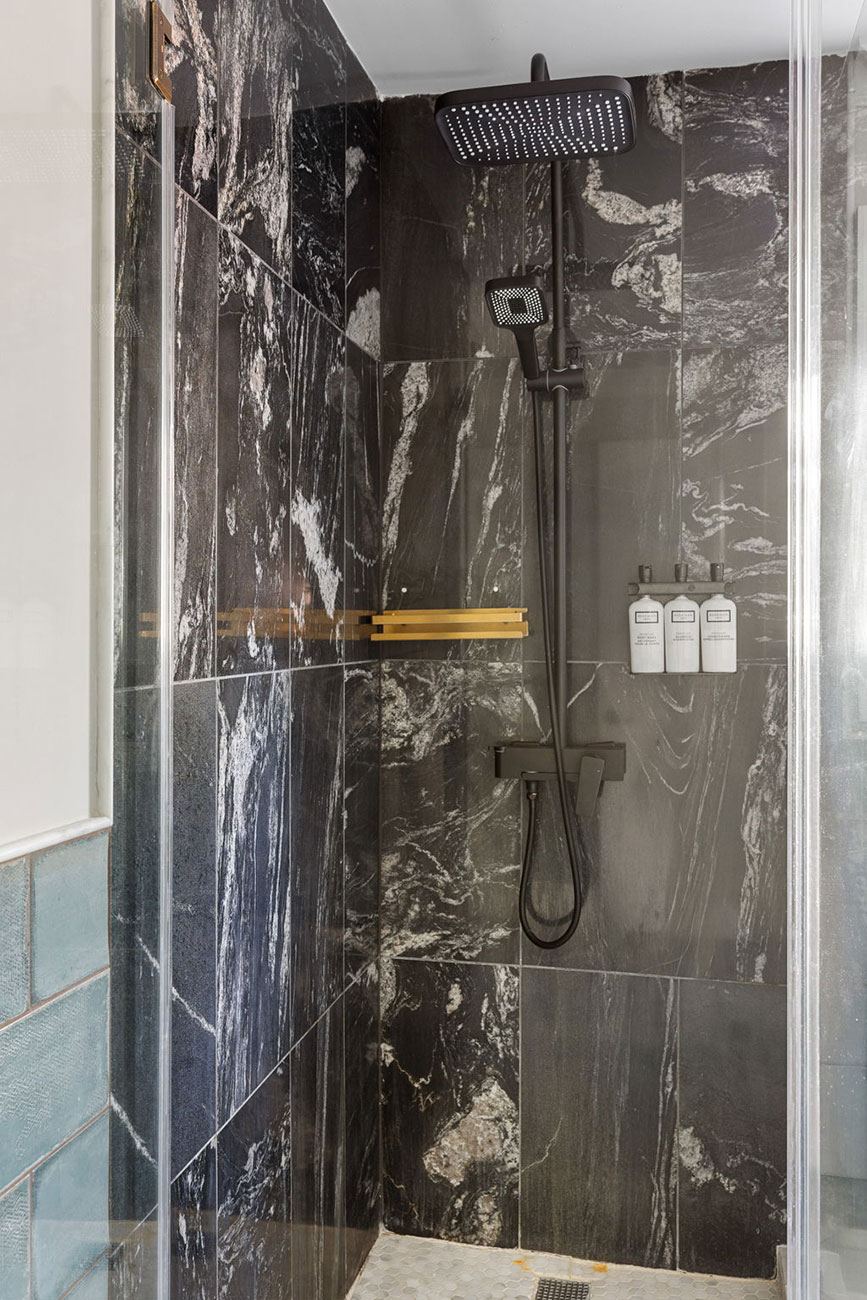 Inside a glass-walled shower with dark marble tiles, a square rainfall head, a handheld wand, a gold towel rack, and a shelf with white bottles.