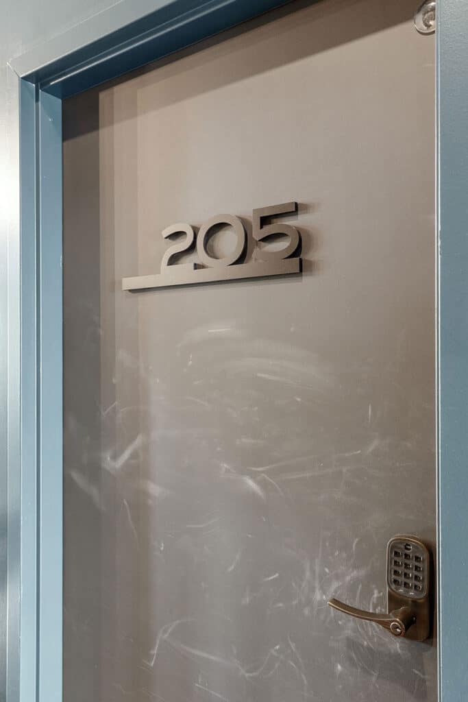 Metal door with the number 205 mounted above a horizontal bar; blue door frame and a bronze keypad lock beside a lever handle.