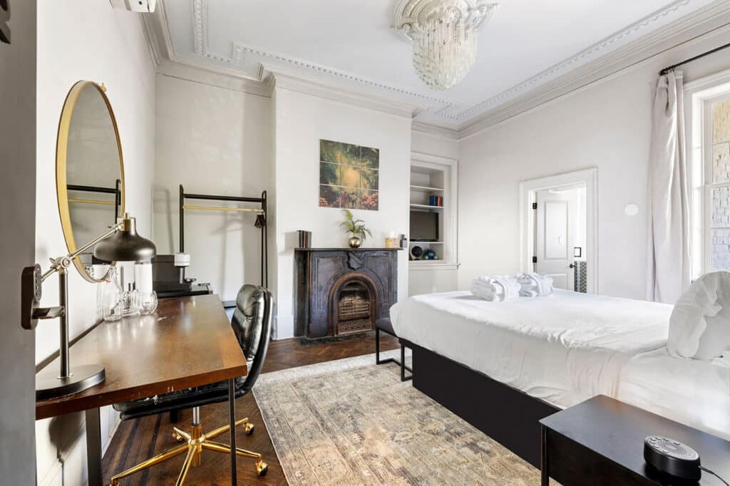 Hotel bedroom with a writing desk, leather chair, round mirror, antique fireplace, and a neatly made white bed.