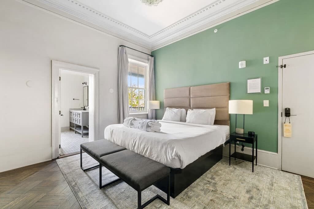 Hotel room with a white bed and tan upholstered headboard, green accent wall, and a bench at the foot near an open bathroom doorway.