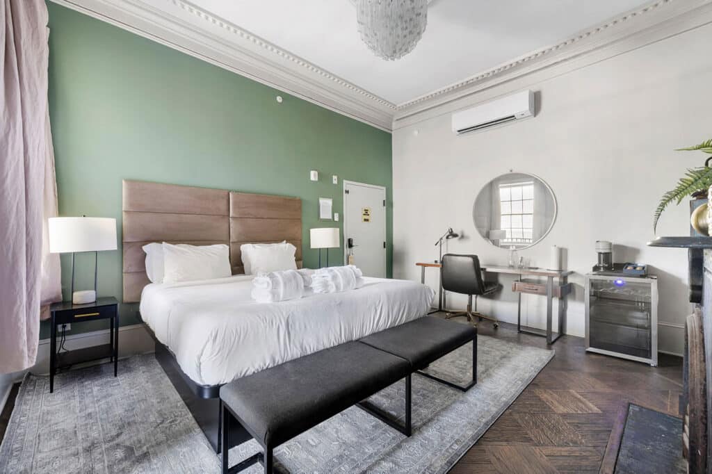 Hotel room with a king bed, white linens, and a green accent wall; bench at foot, bedside lamps, and a desk area with round mirror and minibar.