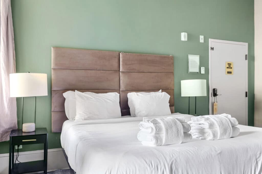 Hotel bedroom with a large padded headboard, white bedding, and folded towels on the bed against a green wall; two bedside lamps nearby.