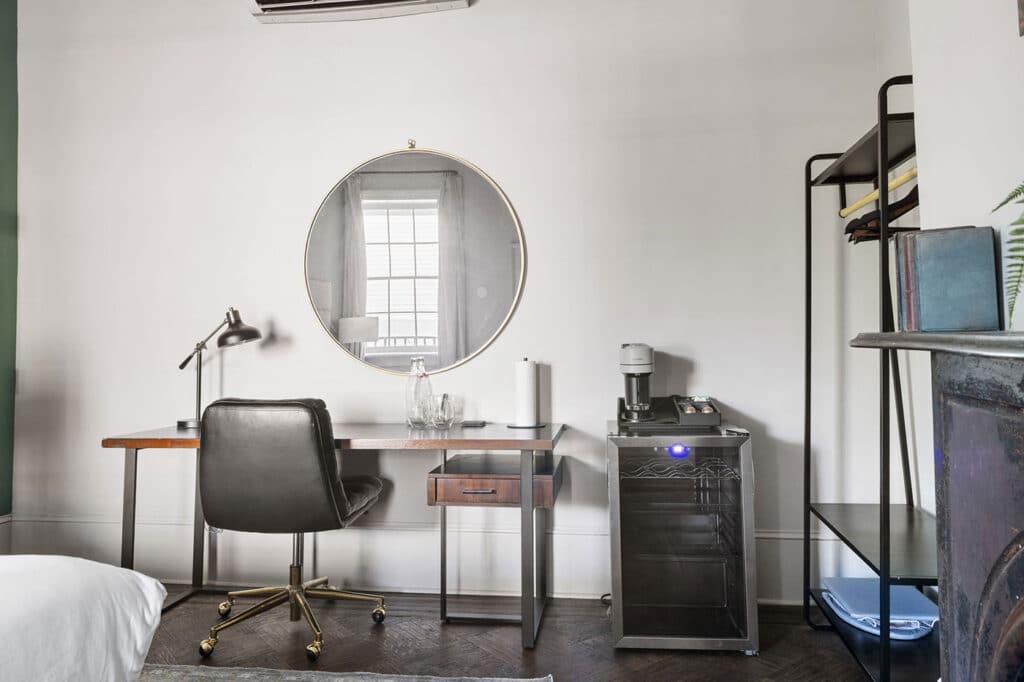 Modern bedroom workspace: round mirror above a wooden desk, black swivel chair, and a small beverage station with a coffee maker to the right