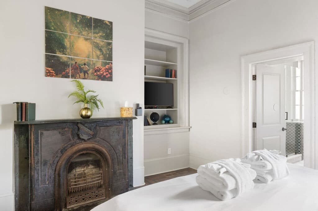 Bright bedroom with a dark ornate fireplace, a potted plant on the mantel, wall art, built-in shelves with a TV, and folded towels on the bed.