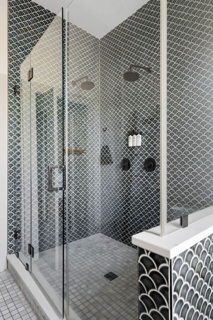 Glass-enclosed shower with monochrome scallop-pattern tiles, two rainfall shower heads on opposite walls, a corner shelf with bottles, and a glass door with metal hinges.