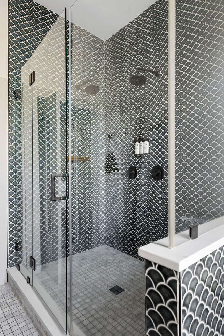 Glass-enclosed shower with monochrome scallop-pattern tiles, two rainfall shower heads on opposite walls, a corner shelf with bottles, and a glass door with metal hinges.