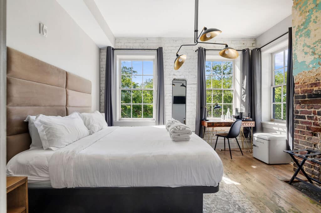Bright bedroom with white bedding, beige tufted headboard, white brick wall, and large windows with gray curtains.