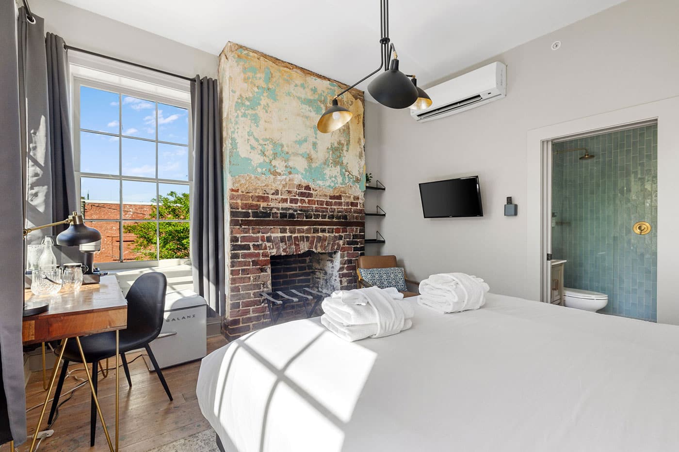Bright hotel room with a brick fireplace wall, large window, desk by the window, and a neatly made white bed in the foreground.