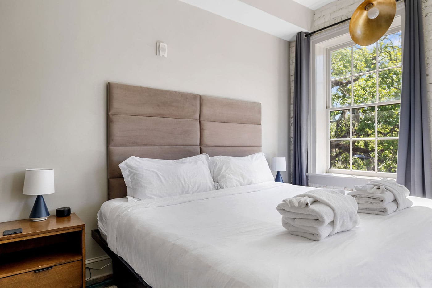 Bright bedroom with a large bed, white linens, and a padded beige headboard; folded towels sit on the bed near the window.
