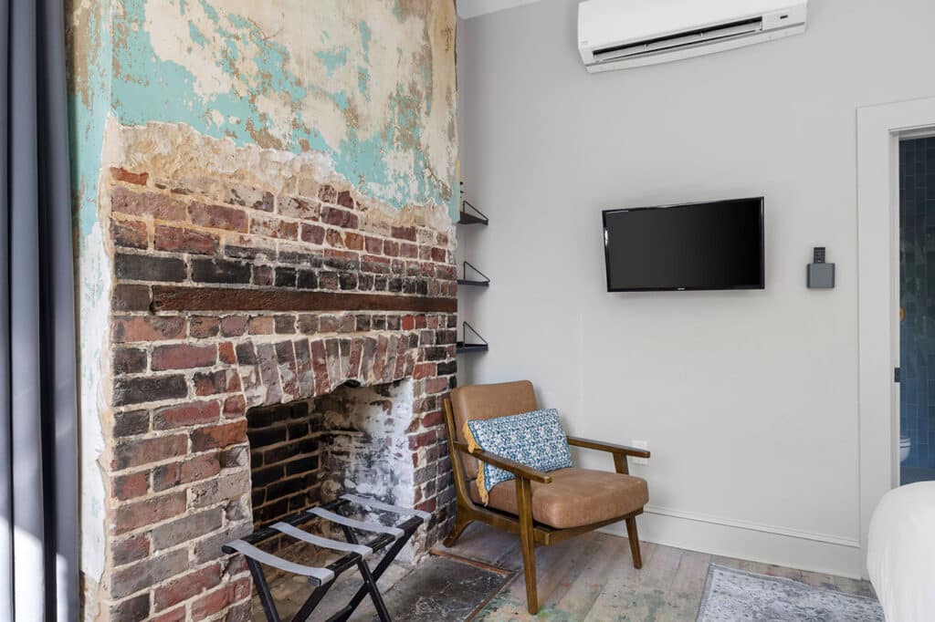Living room with an exposed brick fireplace, peeling turquoise paint, and a mid-century brown armchair with a blue patterned cushion near a wall-mounted TV.