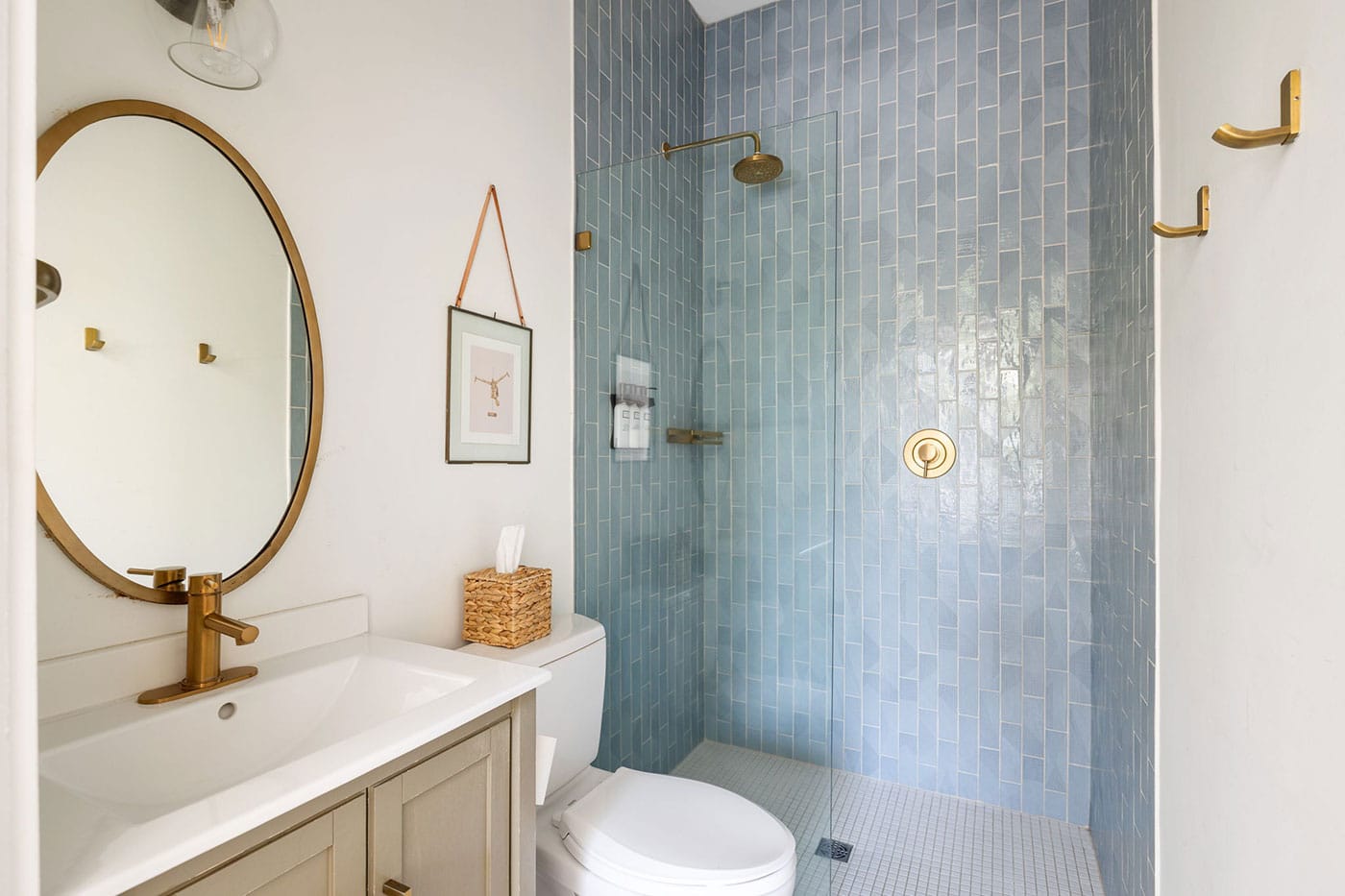 Blue-tiled walk-in shower with brass fixtures beside a white vanity and round gold-framed mirror in a modern bathroom with a toilet nearby.