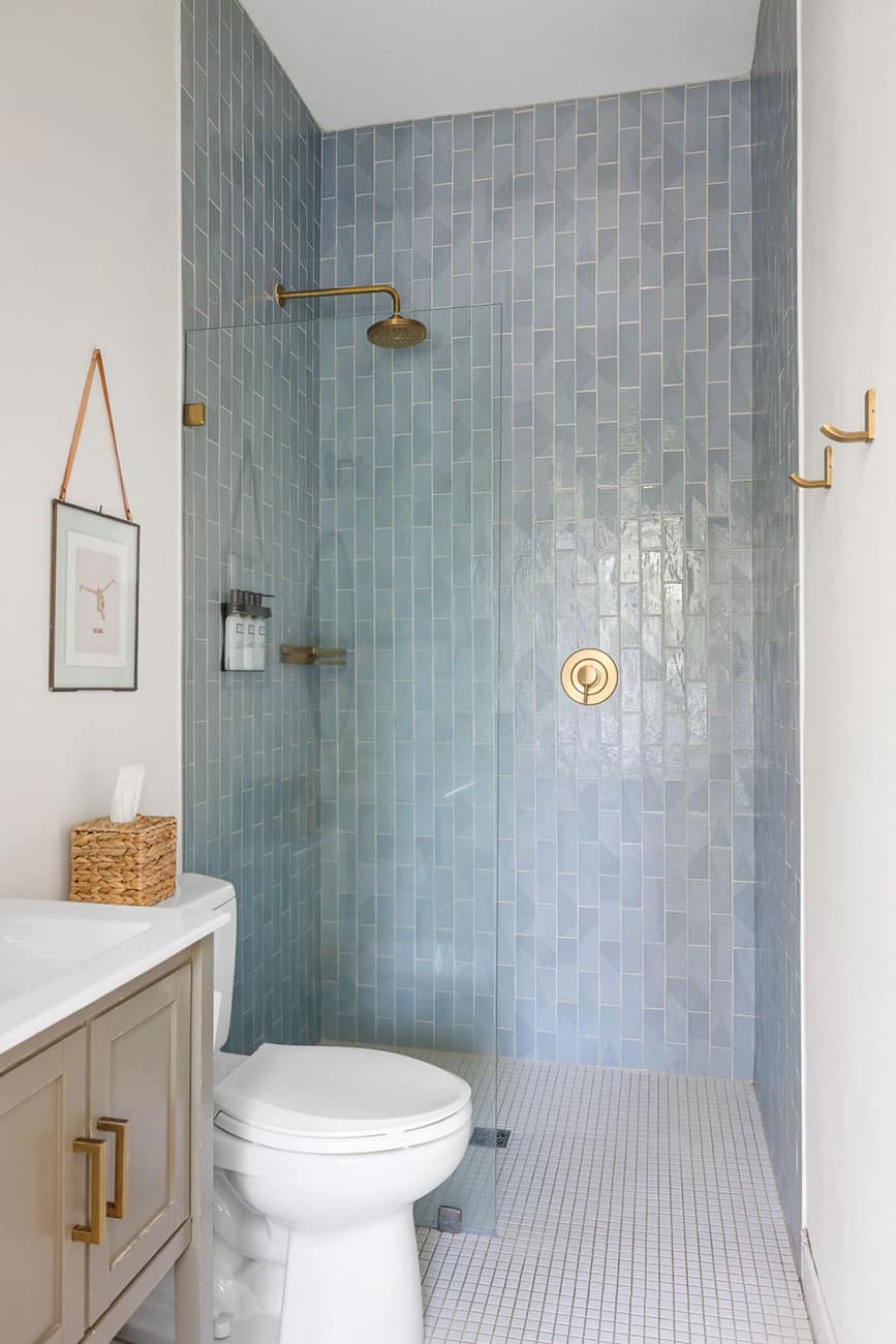 Blue-tiled shower with brass fixtures and a clear glass panel beside a white toilet and beige vanity.