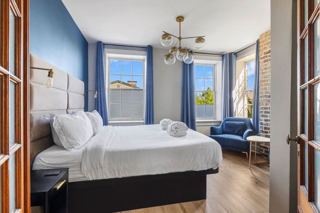 Bright bedroom with a white bed, blue accent wall, and twin blue curtains; a blue chair sits by a brick column and a small table nearby.