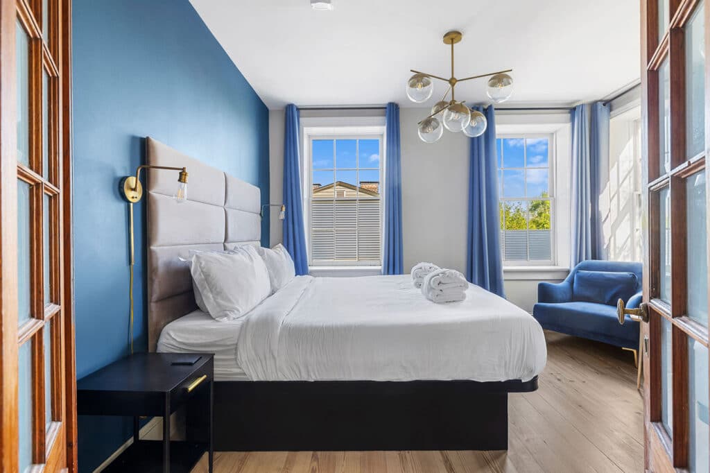 Hotel bedroom with a blue accent wall, upholstered headboard, white bedding, and a blue armchair by the windows.