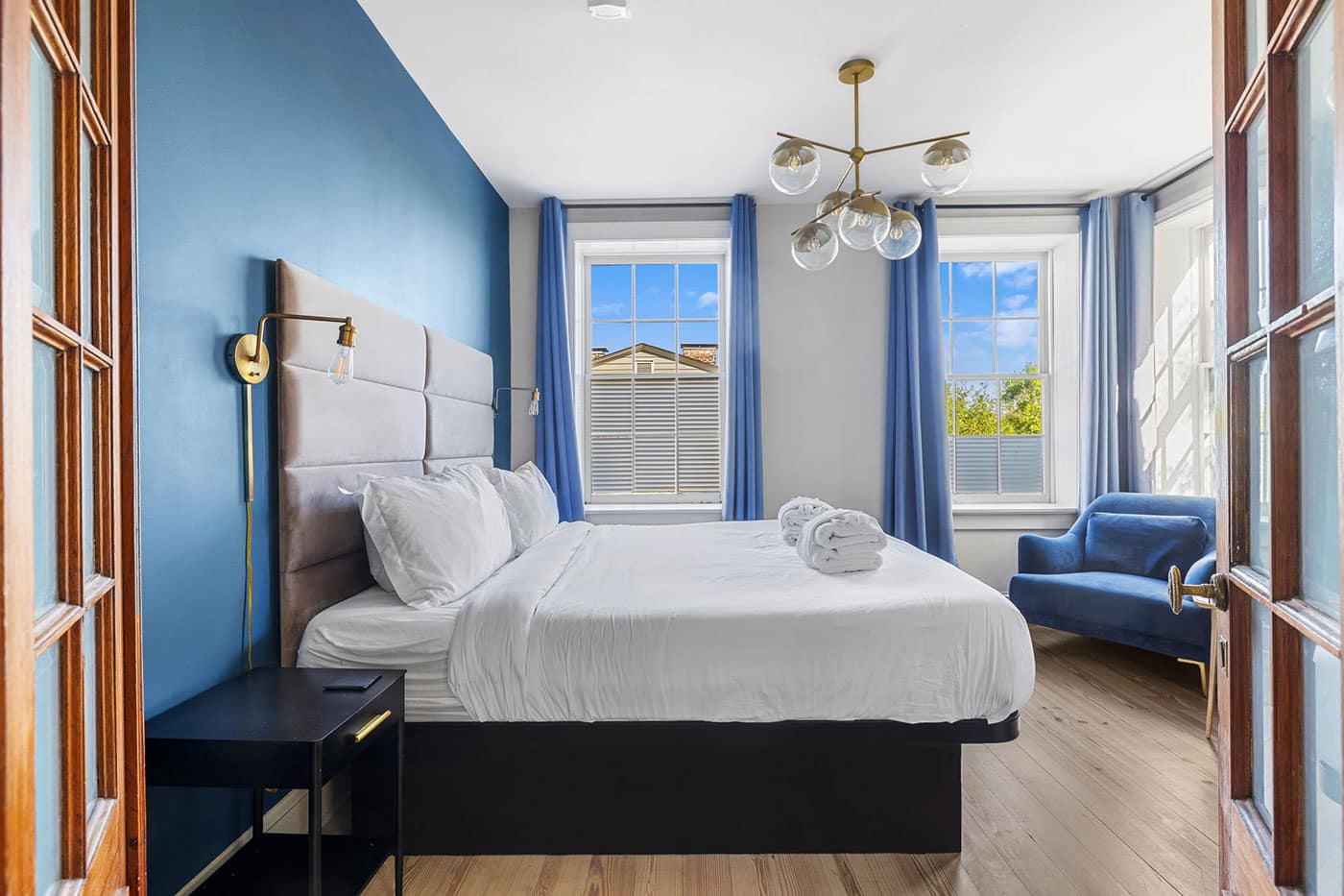 Hotel bedroom with a blue accent wall, upholstered headboard, white bedding, and a blue armchair by the windows.