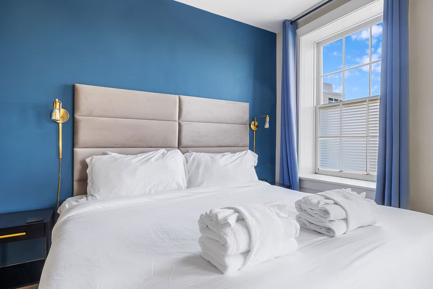Hotel-style bedroom with a blue accent wall, beige upholstered headboard, gold wall lamps, and a white bed with folded towels on top.