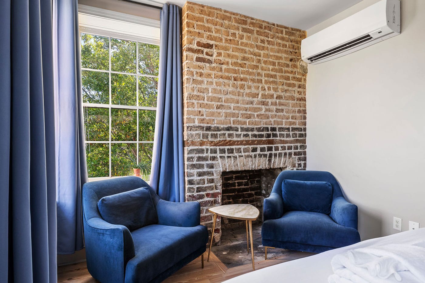 Cozy seating area with an exposed brick fireplace, two blue armchairs, and a small round table near a large window with blue curtains.