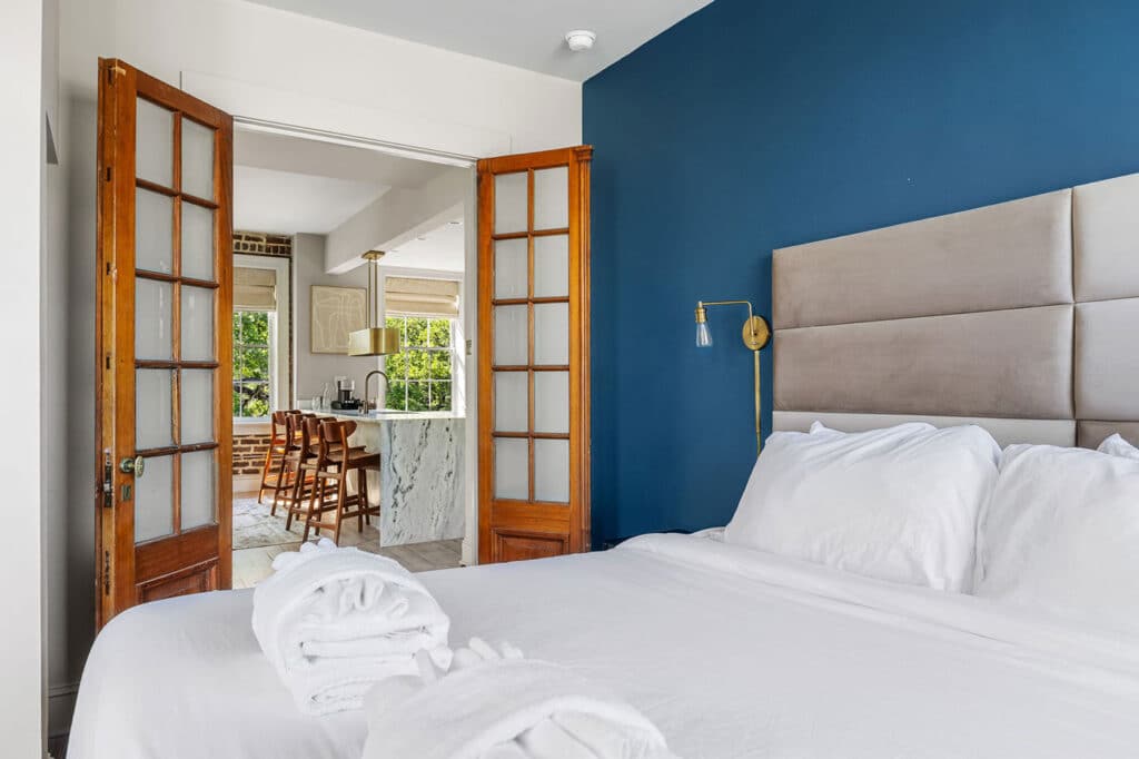 Bedroom with blue accent wall, beige headboard, and white bedding; French doors open to a bright kitchen with a marble island.