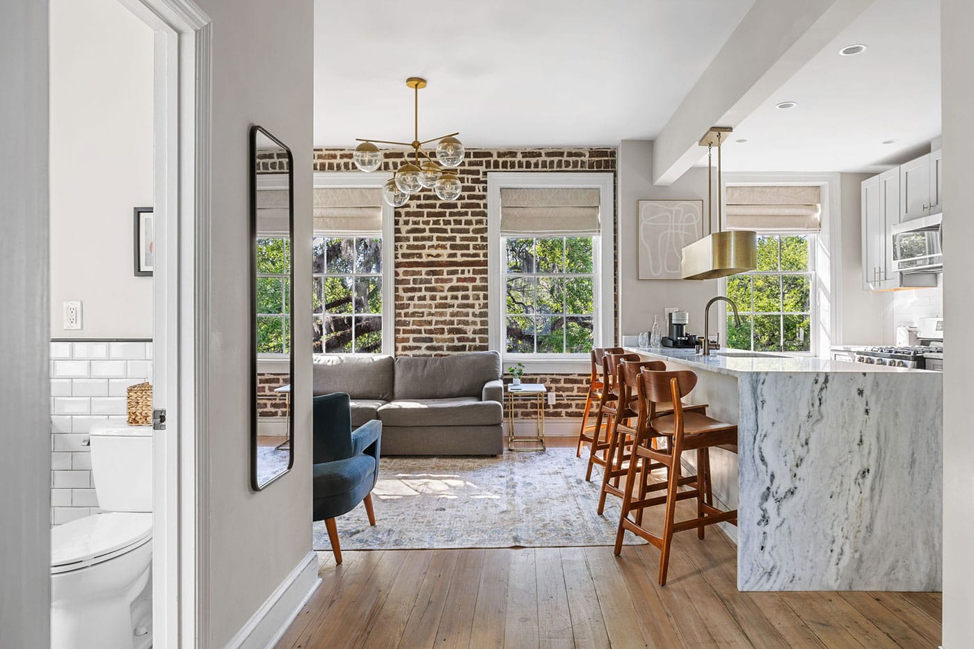 Open-plan living area with an exposed brick wall, large windows, gray sofa, blue accent chair, and a marble kitchen island with wooden stools.