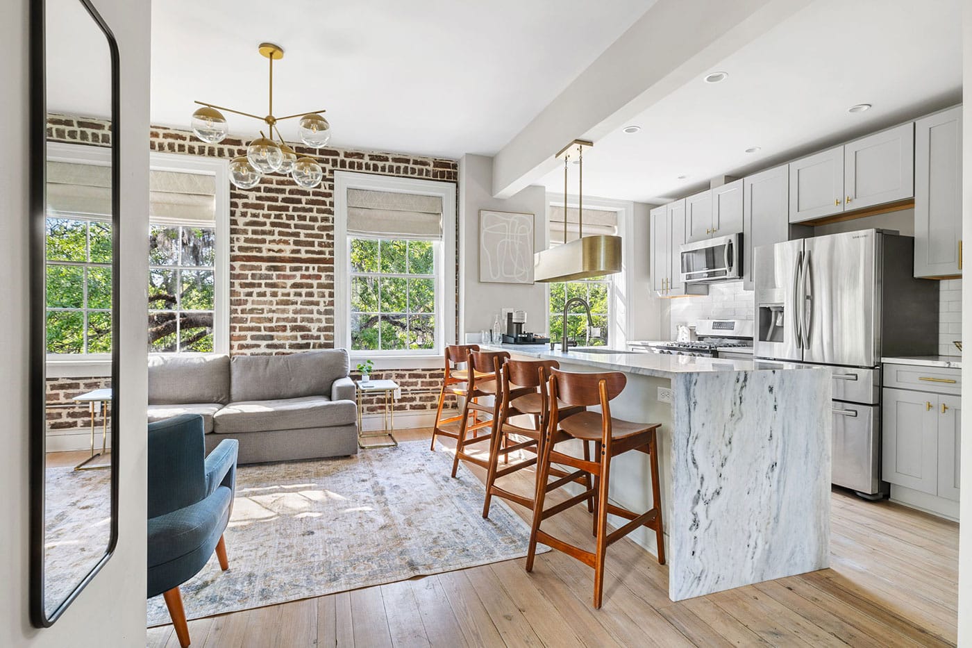 Open-concept living room and kitchen with an exposed brick wall, gray sofa, blue chair, and marble-topped island.