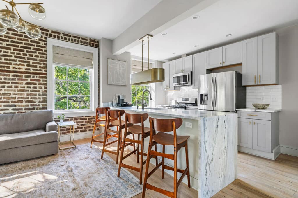 Open-concept kitchen with a marble island, gray cabinets, stainless steel fridge, and four wooden bar stools under a brass pendant light.
