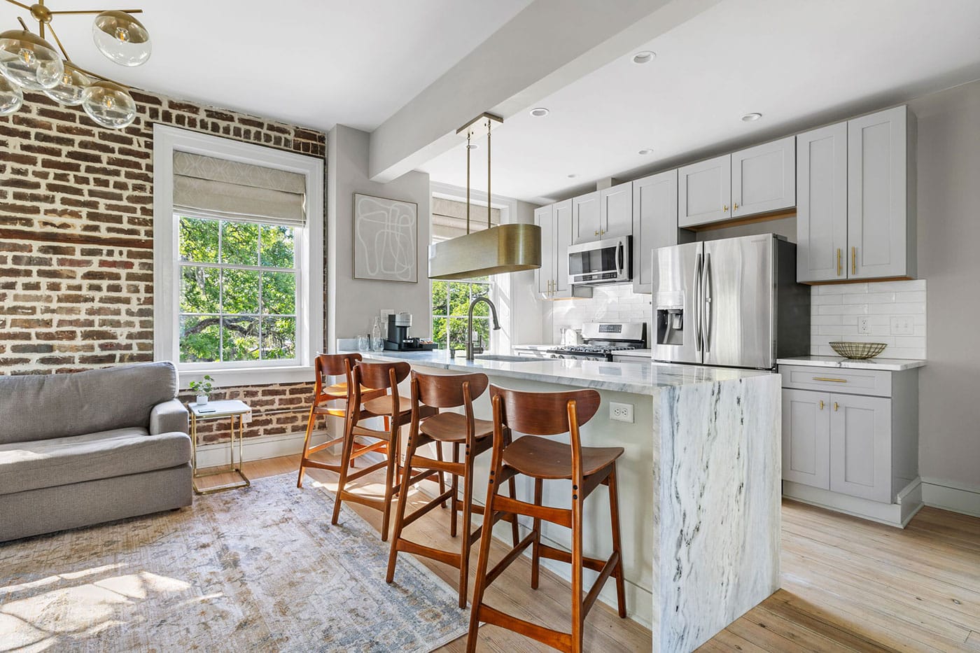Open-concept kitchen with a marble island, gray cabinets, stainless steel fridge, and four wooden bar stools under a brass pendant light.
