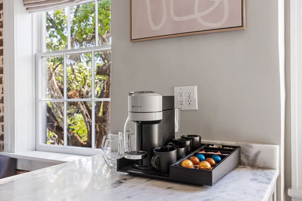Coffee station on a marble countertop with a silver Nespresso machine, mugs, and a tray of colorful capsules beside a window.