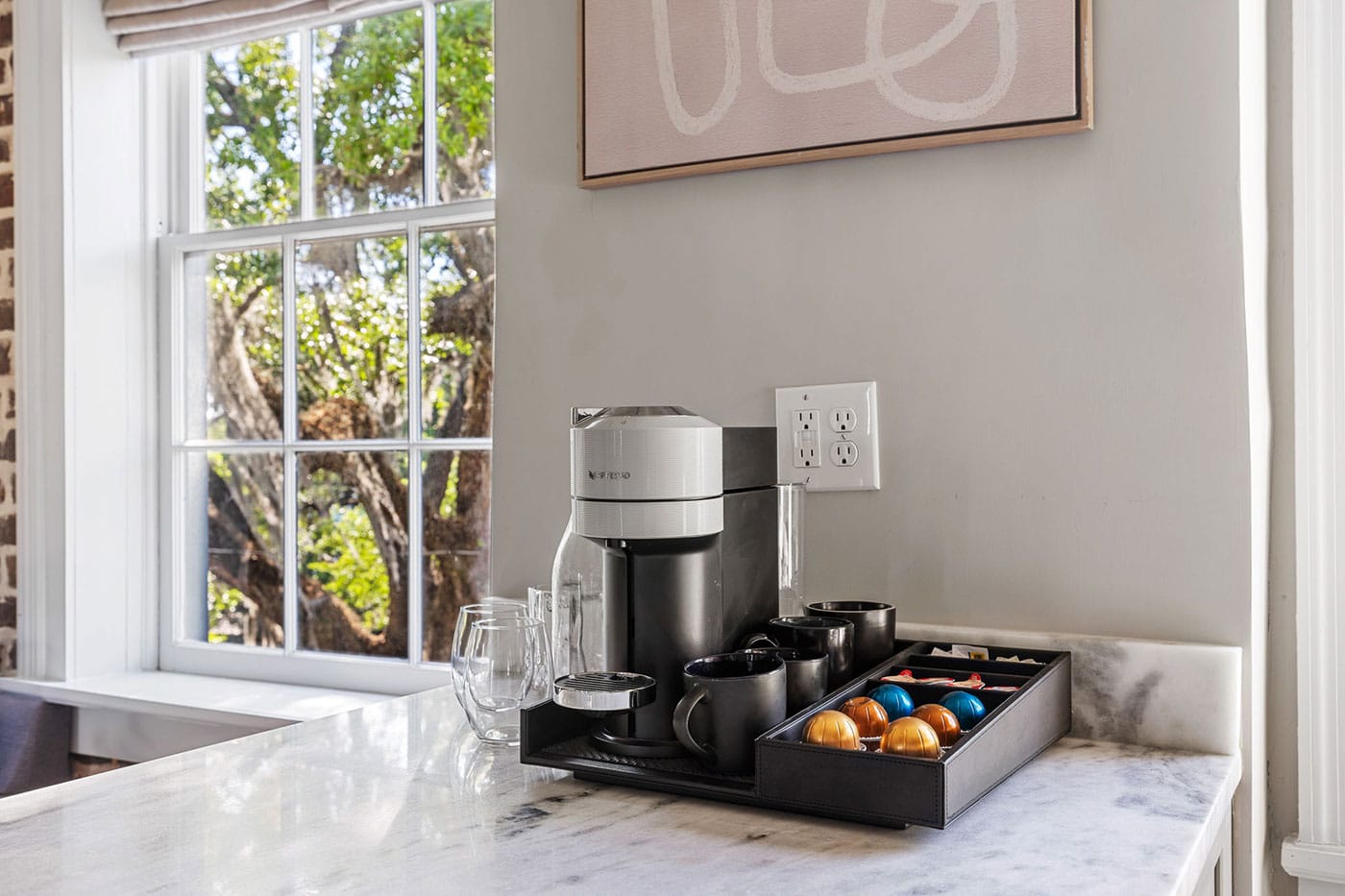 Coffee station on a marble countertop with a silver Nespresso machine, mugs, and a tray of colorful capsules beside a window.