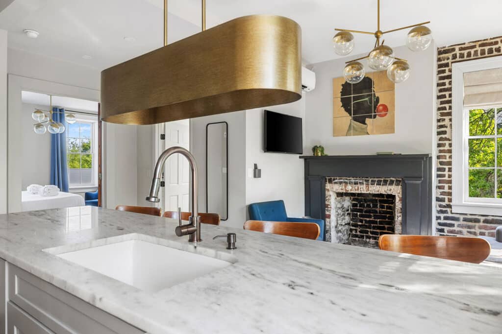 Open-concept kitchen with a marble island and bronze faucet, facing a living room with a brick fireplace and modern art above the mantle.