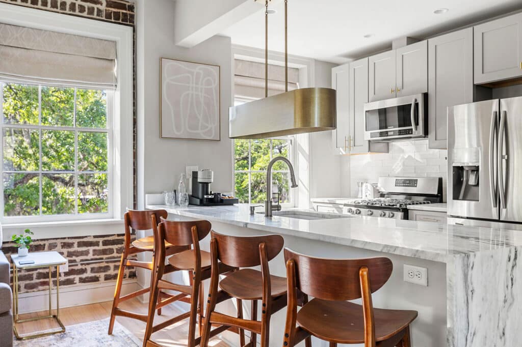 Modern kitchen with marble island, stainless steel appliances, and wooden bar stools by a bright windowed breakfast bar.
