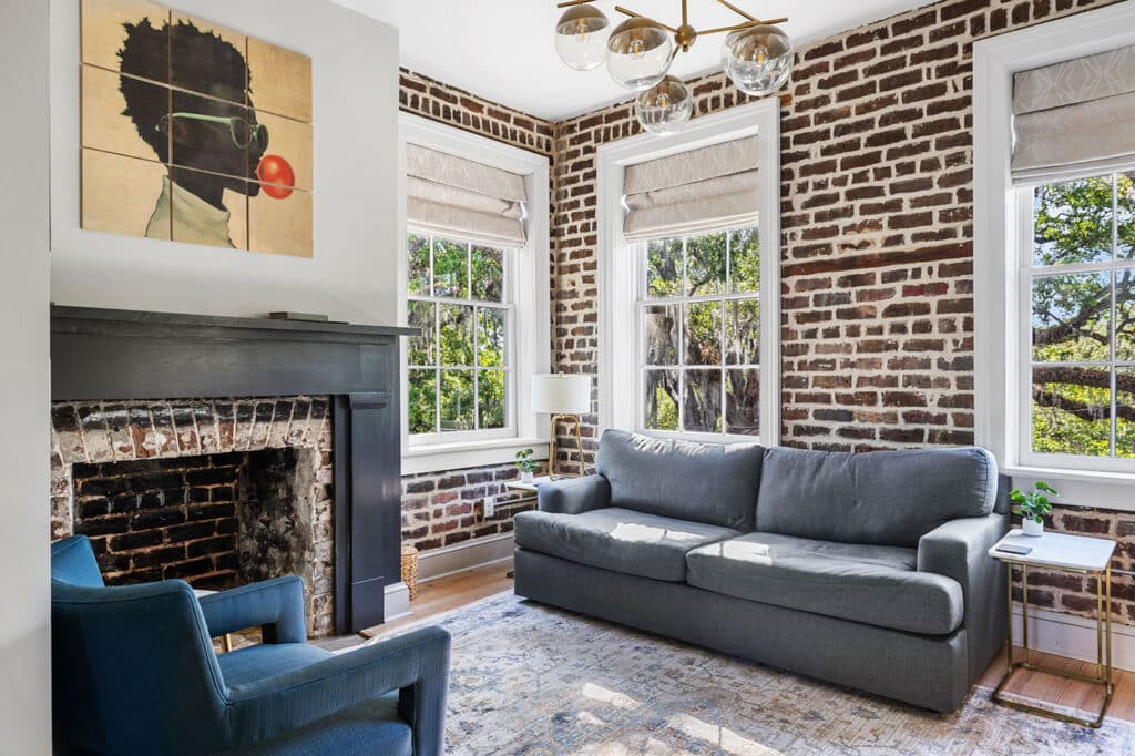 Cozy living room with exposed brick walls, a gray sofa, blue accent chair, and a brick fireplace under a multi-panel portrait. Large windows with Roman shades let in natural light.