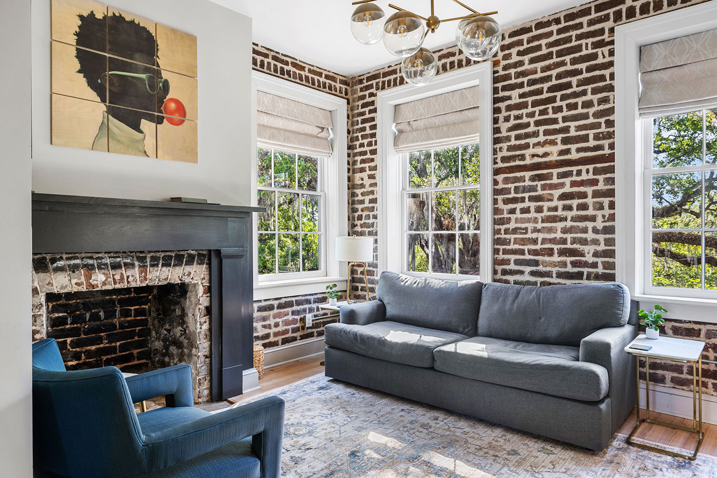 Cozy living room with exposed brick walls, a gray sofa, blue accent chair, and a brick fireplace under a multi-panel portrait. Large windows with Roman shades let in natural light.