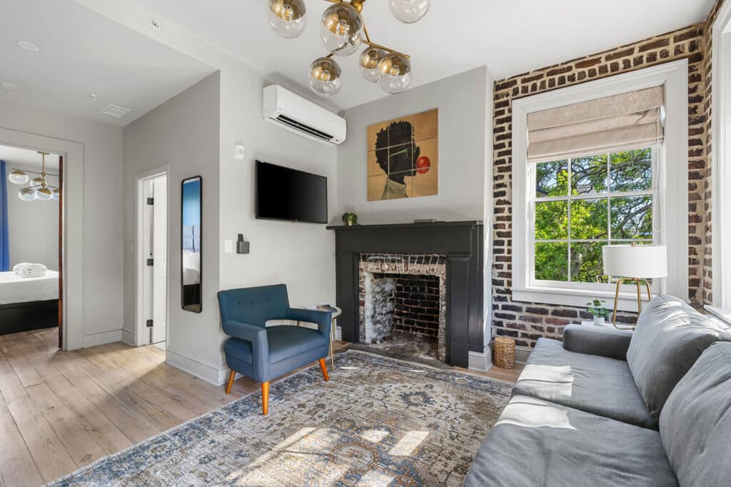 Bright living room with gray walls, black fireplace mantel, wall-mounted TV, blue armchair, and a large patterned rug.